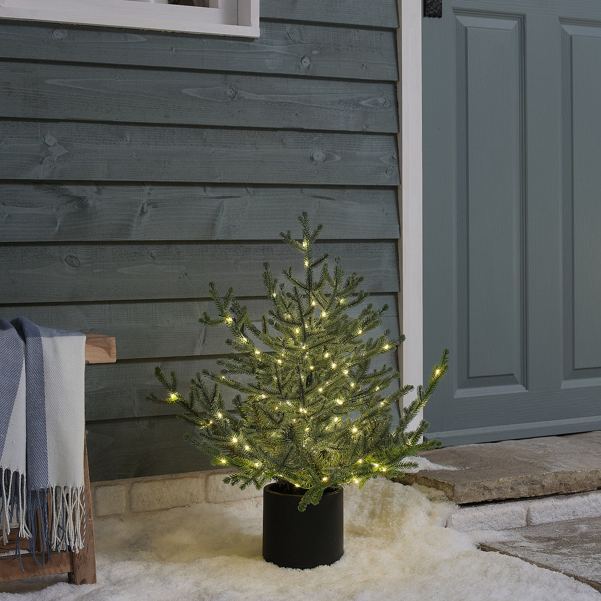 Beleuchteter LED-Weihnachtsbaum im Topf steht draußen vor Hauswand mit Schnee