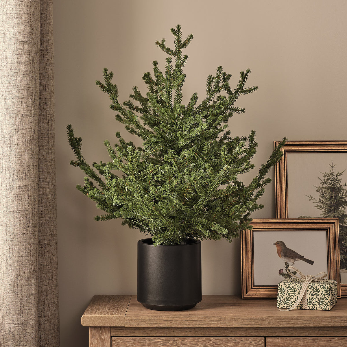 Künstlicher Weihnachtsbaum im Topf ohne Beleuchtung auf Holztisch vor Wand mit Bildern