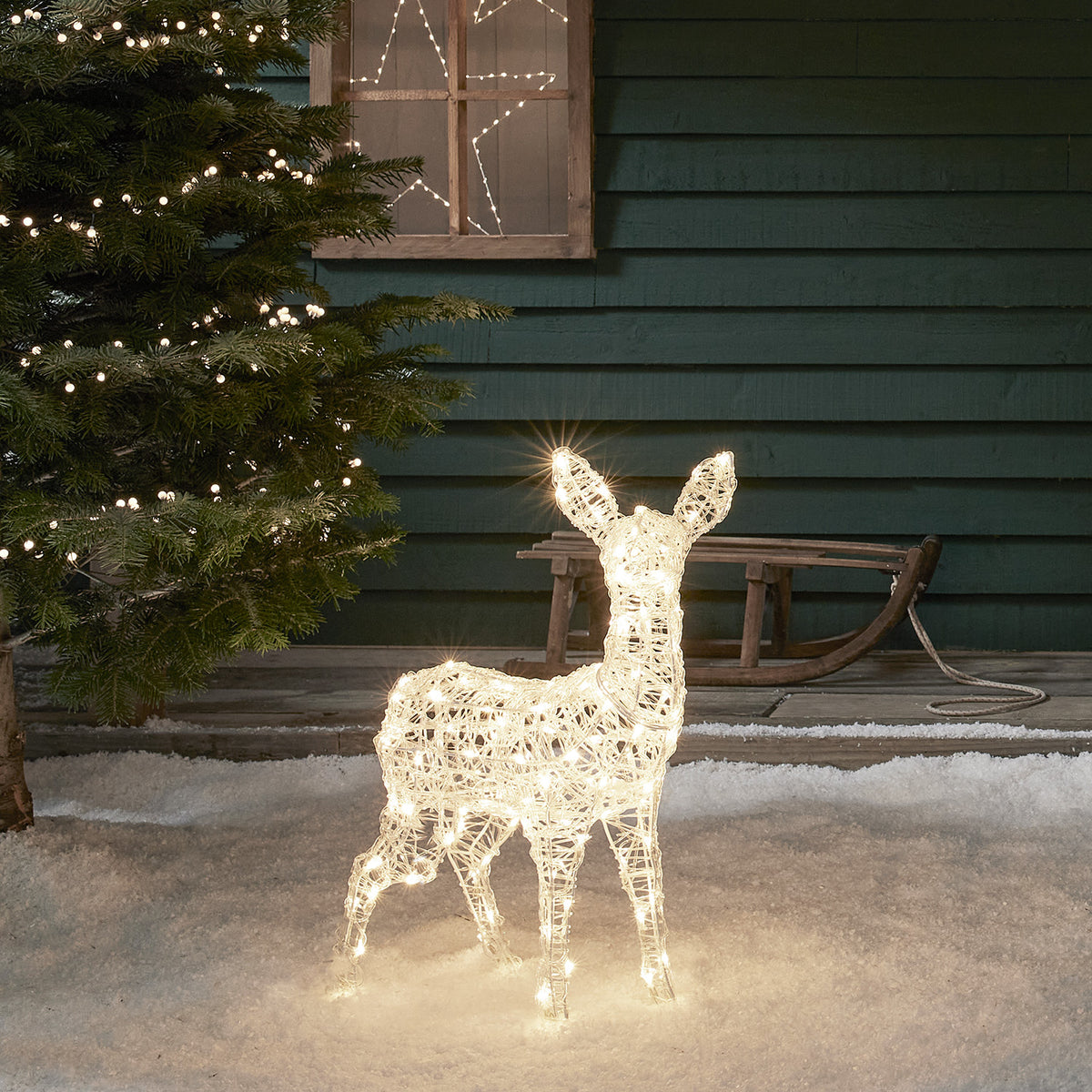 Beleuchtetes Solar-LED-Rehkitz aus Acryl steht im Schnee vor grünem Haus mit Weihnachtsbaum