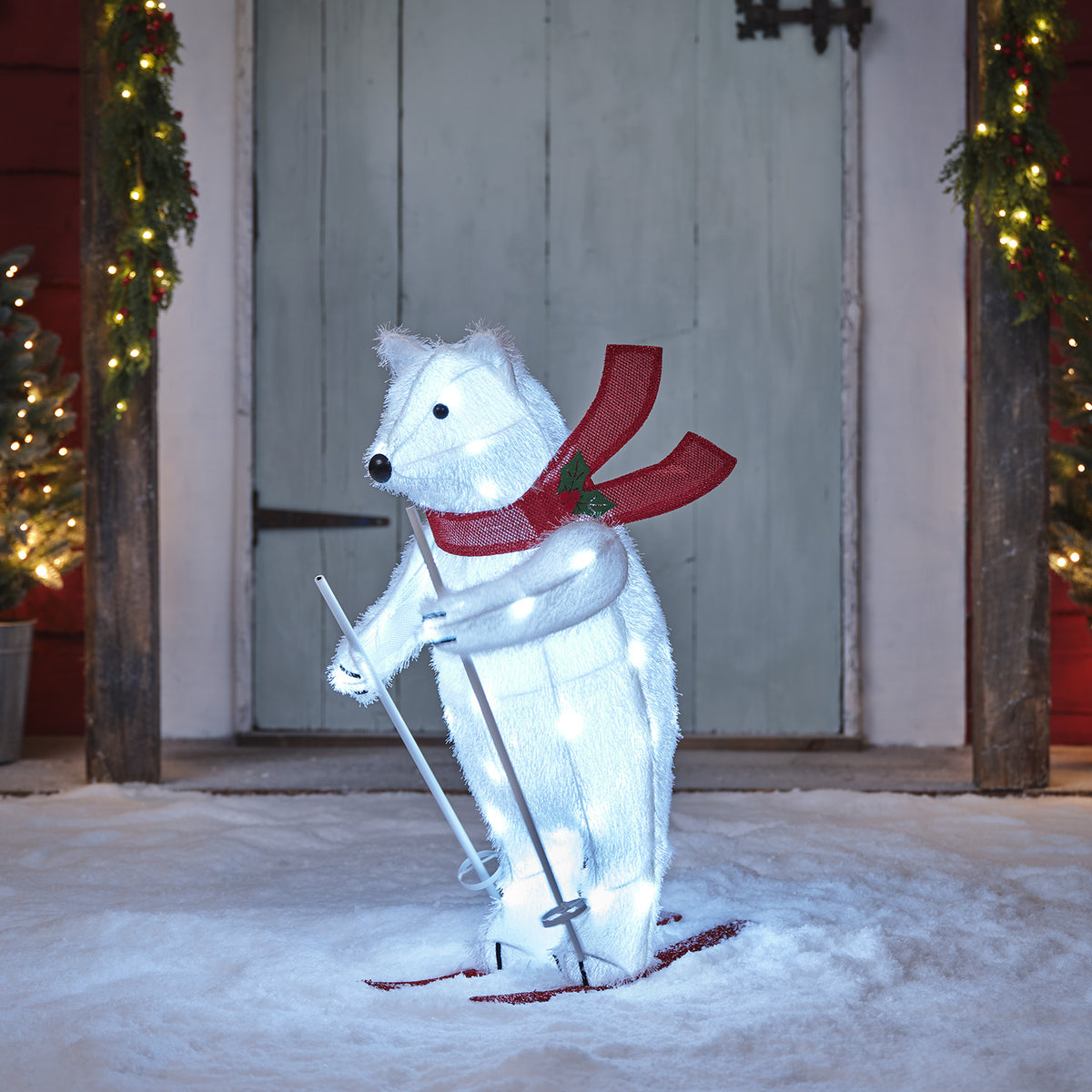 Leuchtende LED-Weihnachtsfigur Polarbär mit rotem Schal und Skiern vor Holztür im Schnee