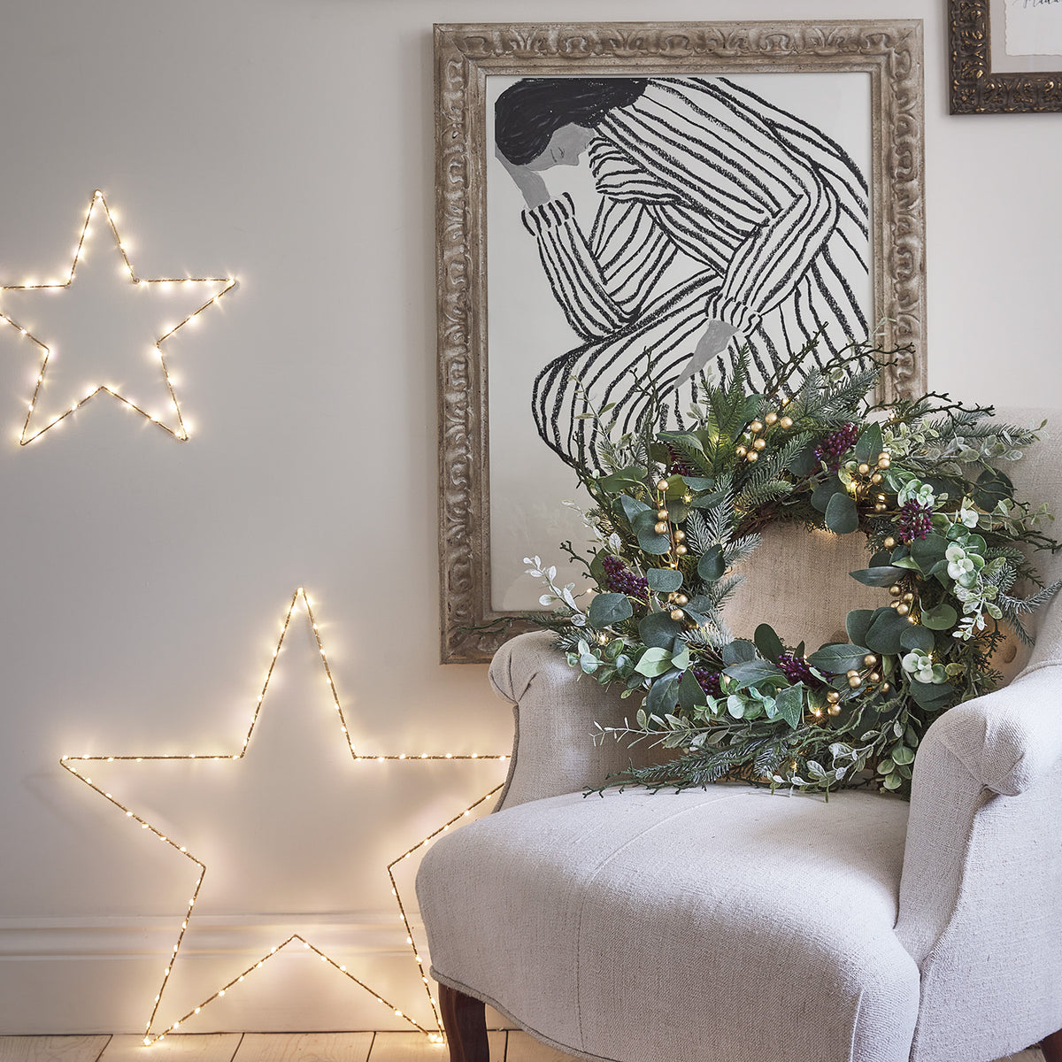 Weihnachtskranz mit Lichterkette auf einem beigen Sessel vor Wand mit Sternenbeleuchtung