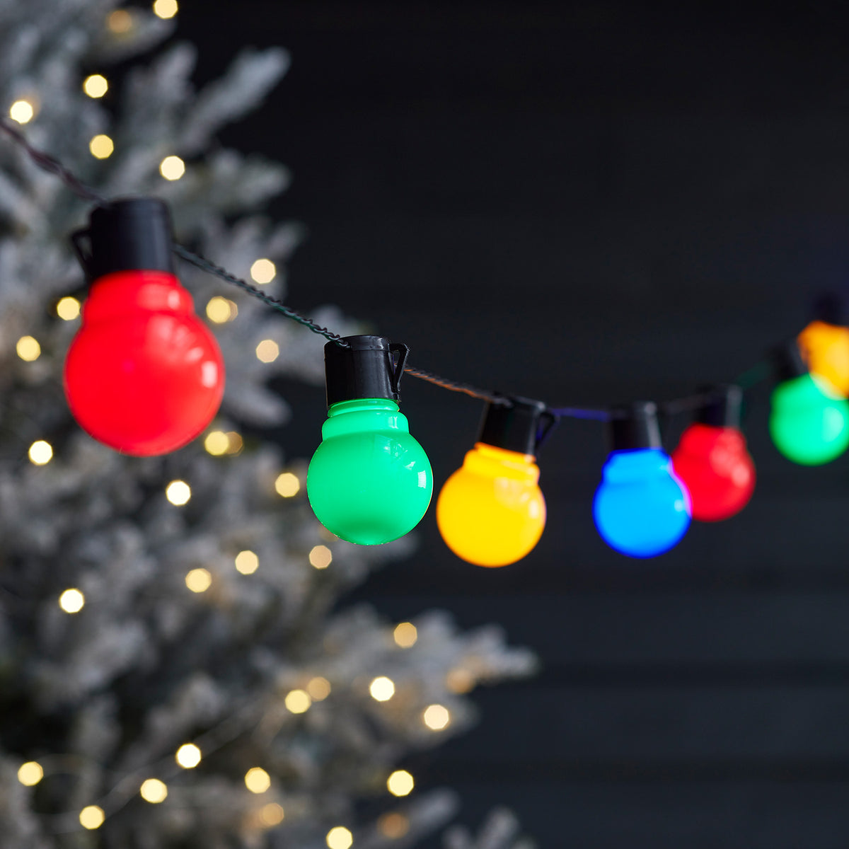 Bunte LED-Kugeln an schwarzer Lichterkette vor dunklem Hintergrund und unscharfem Baum mit Lichtern